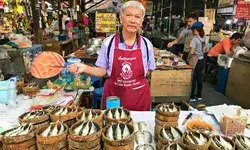 A person standing at a market stall displaying fish in bamboo baskets holding a fan