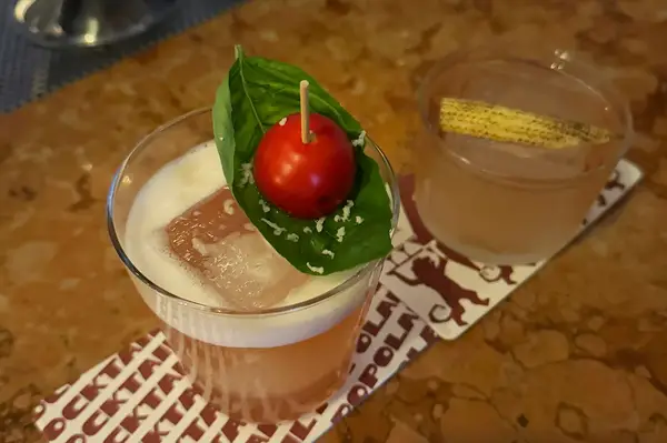 Two crafted cocktails on a table one garnished with a basil leaf and mini tomato the other with a small corn cob