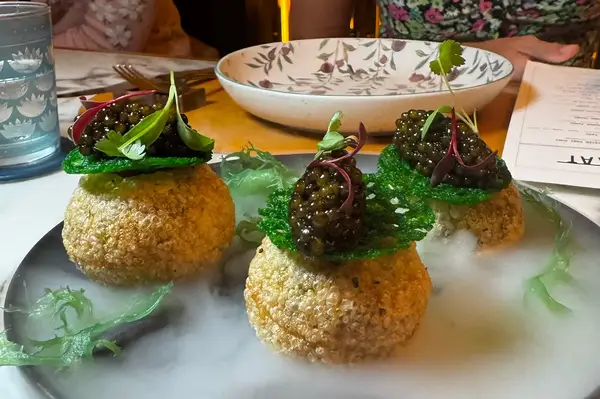 A plate containing three spherical appetizers garnished with caviar and herbs served on a foggy presentation contrasting with decorative tableware in the background