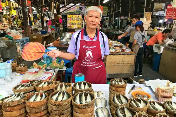 A person standing at a market stall displaying fish in bamboo baskets holding a fan