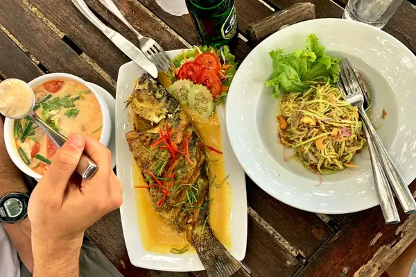 A table setting with plates of Thai food including a whole fried fish a bowl of soup and a salad with a hand holding a spoon