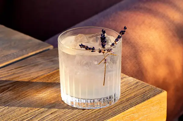A cocktail served in a clear glass with ice and a sprig garnish, placed on a wooden table