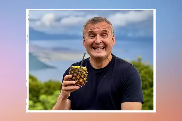 Philip Rosenthal smiling, holding a pineapple drink in an outdoor setting with hills and a body of water in the background
