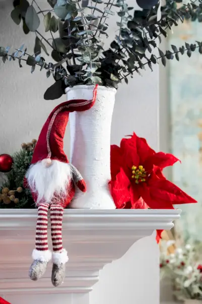 a classic norweigan holiday christmas gnome decoration sitting on a white modern mantle with a white vase with eucalyptus and a poinsettia bloom