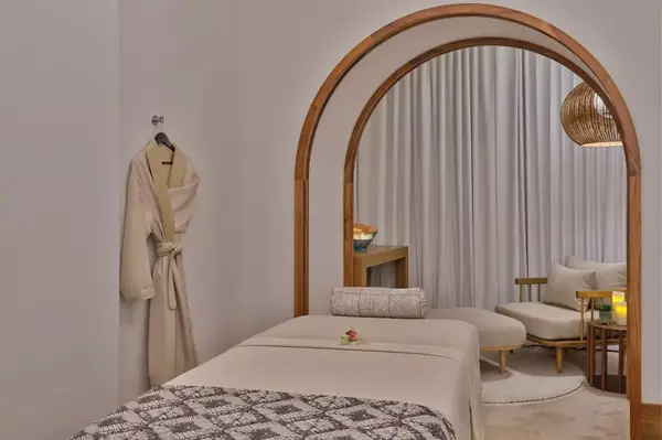 A tranquil spa treatment room featuring a massage table, a robe on a hook, cushioned chairs, and soft lighting