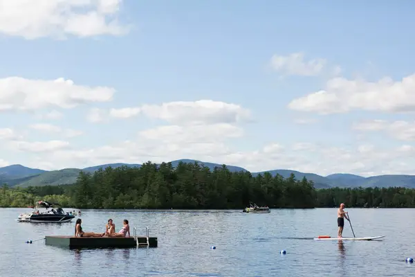 A scenic lakeside view with people paddleboarding and a group on a floating dock, surrounded by distant forested hills
