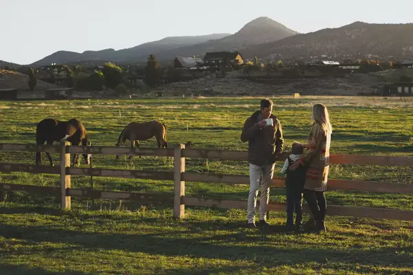 Family standing by a fence in a ranch with horses grazing nearby at a scenic backdrop of hills