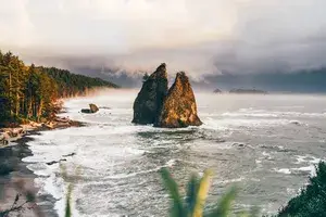 Coastal scene with large rock formations rising out of the ocean, with a sandy beach and forests in the background