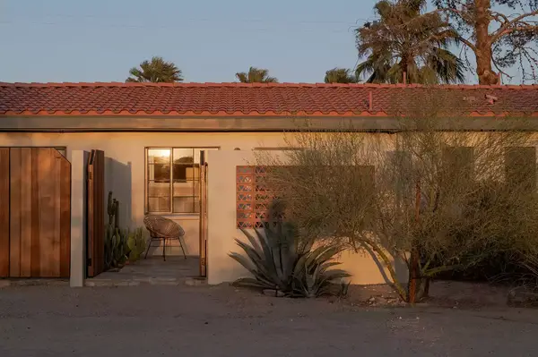 Exterior of a hotel room entrance with a chair visible on the patio and desert plants in the surroundings