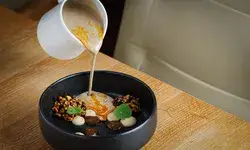 A culinary dish with sauce being poured from a jug into a black bowl containing various garnishes