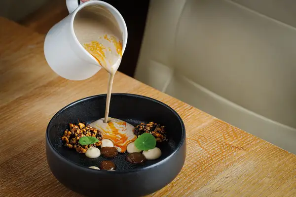 A culinary dish with sauce being poured from a jug into a black bowl containing various garnishes