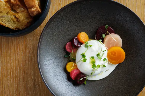 A plate with burrata cheese garnished with assorted vegetables and greens and a side of toasted bread