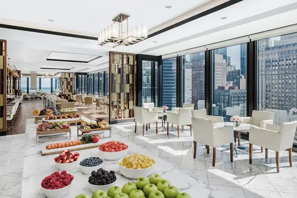A luxurious dining area with large windows overlooking a cityscape tables set with chairs and a buffet with fruits and other items