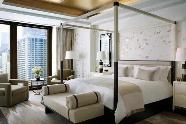 Luxurious hotel room featuring a canopy bed armchairs a bench and large windows with a cityscape view