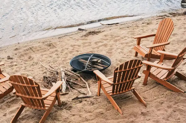 Wooden chairs arranged around a firepit on a sandy lakeside shore