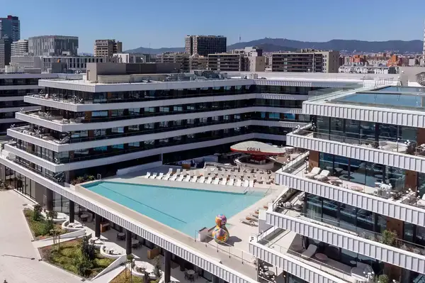 A modern hotel pool area surrounded by a large building complex, located in an urban setting with cityscape in the background
