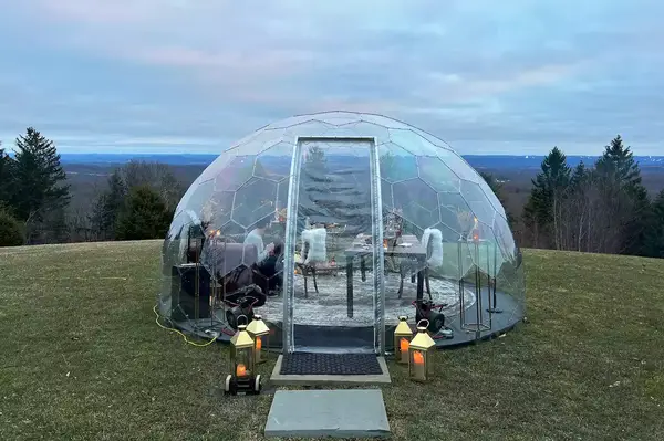 A transparent igloo dining setup on a grass field with a scenic landscape view in the background, decorated with lanterns