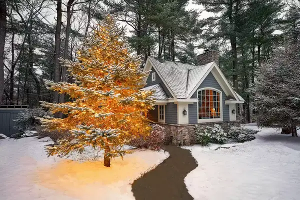Snow-covered guest cottage in a forested area with a lit tree in the foreground, winter setting