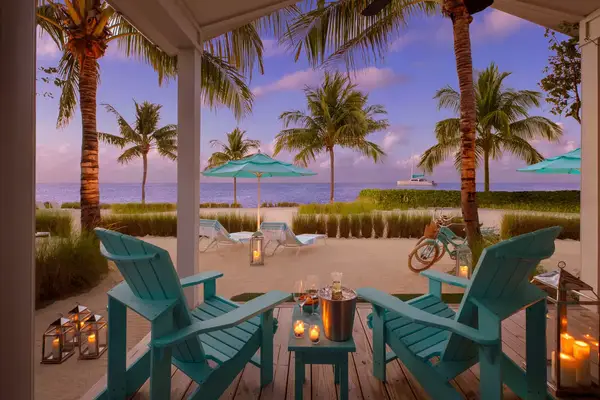 Patio with chairs and table overlooking a beach, palm trees, umbrellas, and lanterns in view.