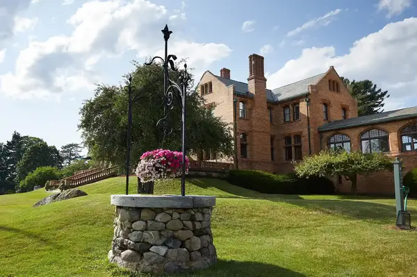 A historic brick building with a stone well in front, surrounded by greenery and trees