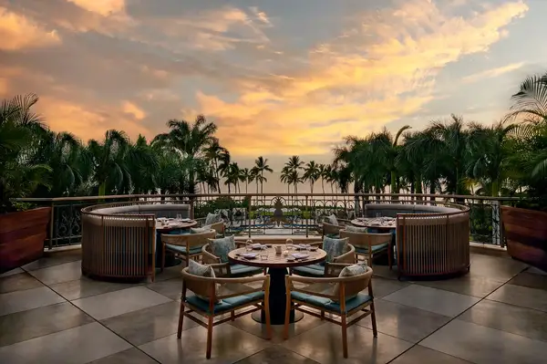Outdoor dining area with round tables and chairs overlooking palm trees and an ocean sunset