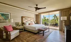 Interior of a hotel suite with a bed, seating area, balcony, and tropical outdoor view