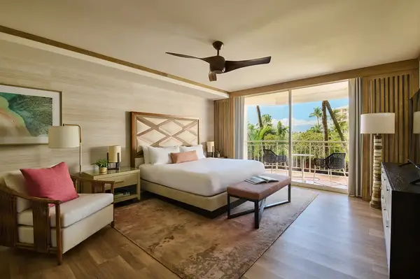 Interior of a hotel suite with a bed, seating area, balcony, and tropical outdoor view