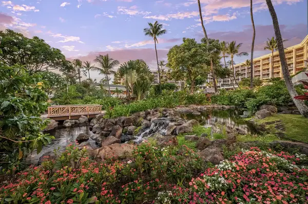 resort garden featuring a landscaped stream bordered by flowers and trees, surrounded by resort buildings and tropical scenery