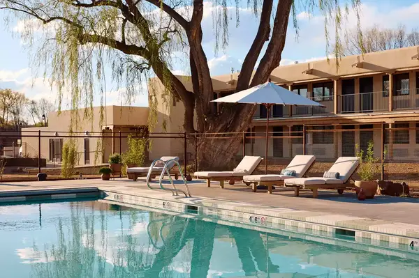 Outdoor pool area with lounge chairs, umbrella, and a tree near a beige building