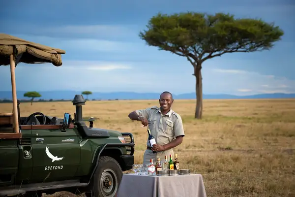 A person standing beside a safari vehicle in a grassy plain setting preparing a drink at a small table