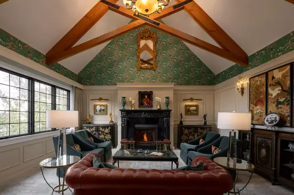 Elegant living room with exposed wooden beams, large windows, a patterned wallpaper, and a central fireplace surrounded by seating