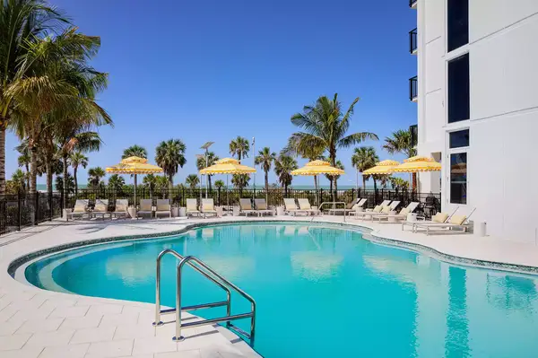 Pool area with lounge chairs and umbrellas by a hotel, palm trees and blue sky in the background