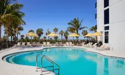 Pool area with lounge chairs and umbrellas by a hotel, palm trees and blue sky in the background