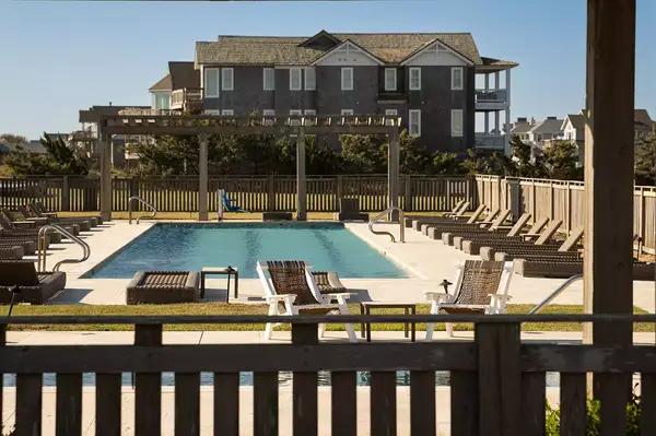 Outdoor pool area at a resort with lounge chairs, a fenced perimeter, and buildings in the background