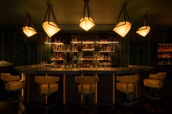 Interior view of a bar with a modern design and hanging lights, featuring a set of bar stools and a backlit shelf with bottles