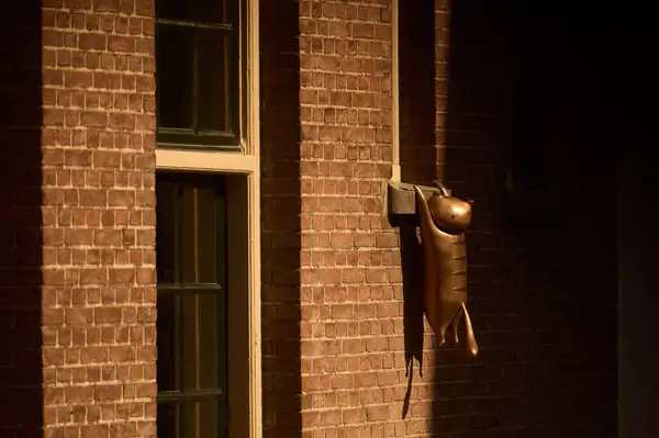 A brick wall with a window, features a bronze sculpture of a hanging figure extending from the wall