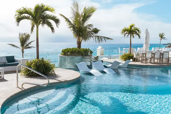 Lounge chairs in a pool on a terrace overlooking the ocean with palm trees and seating areas around