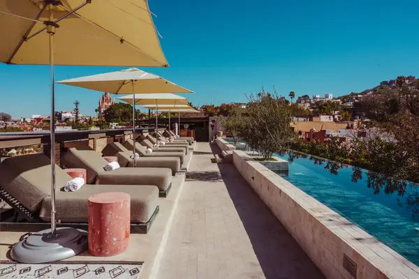 Outdoor lounge area with cushioned chairs, umbrellas, and a pool beside a view of rooftops and trees