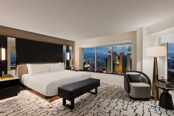 A modern hotel room with a large bed a bench seating a view of MGMs skyline through a window and decorative lighting