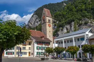 A scenic town square featuring a tall church tower with a clock, surrounded by buildings and greenery, with mountains in the background
