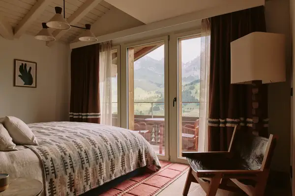 A bedroom with a large window overlooking a balcony and mountainous landscape, featuring a bed, chair, and modern decor
