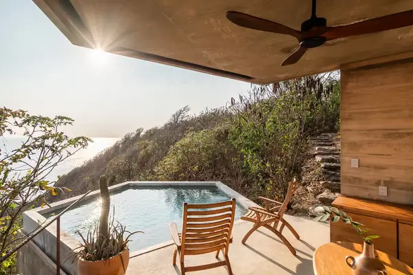 A poolside patio with wooden furniture overlooking hilly vegetation and distant water during sunrise