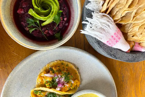 Plated food including two tacos with garnishes a dip in a bowl and a serving of flatbreads with a napkin in a basket
