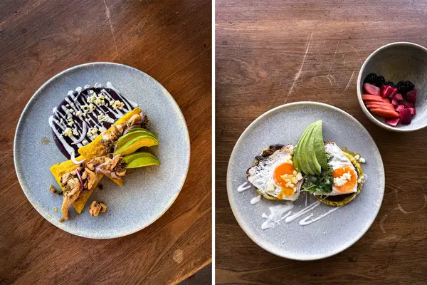 A display of plated dishes including tostadas with eggs and avocado and tacos served on a wooden table with a side of fruit