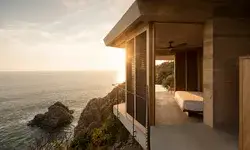 Oceanfacing bedroom with an open balcony featuring coastal rocks in the background and warm natural light from a sunset