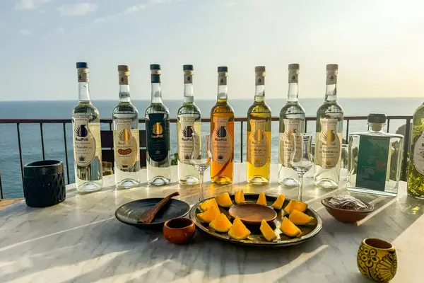 Array of tequila bottles with garnished dishes and glasses arranged on a table against an ocean backdrop
