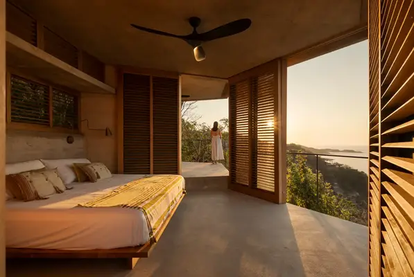 A scenic bedroom with a large bed open wooden shutters and a person in the distance standing on a balcony overlooking nature