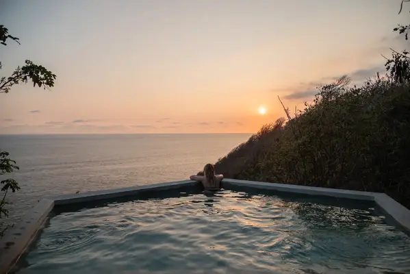 Person leaning on the edge of an infinity pool overlooking a sunset ocean view with surrounding hills