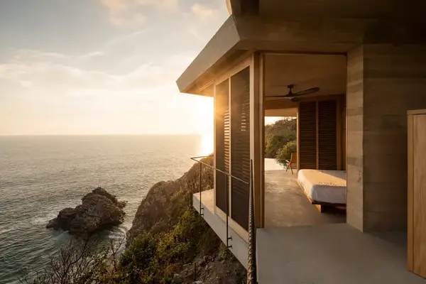 Oceanfacing bedroom with an open balcony featuring coastal rocks in the background and warm natural light from a sunset