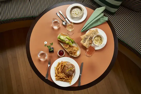 A round table with assorted plated dishes, including a sandwich, soup, a burger with fries, and bread, set with drinks and utensils
